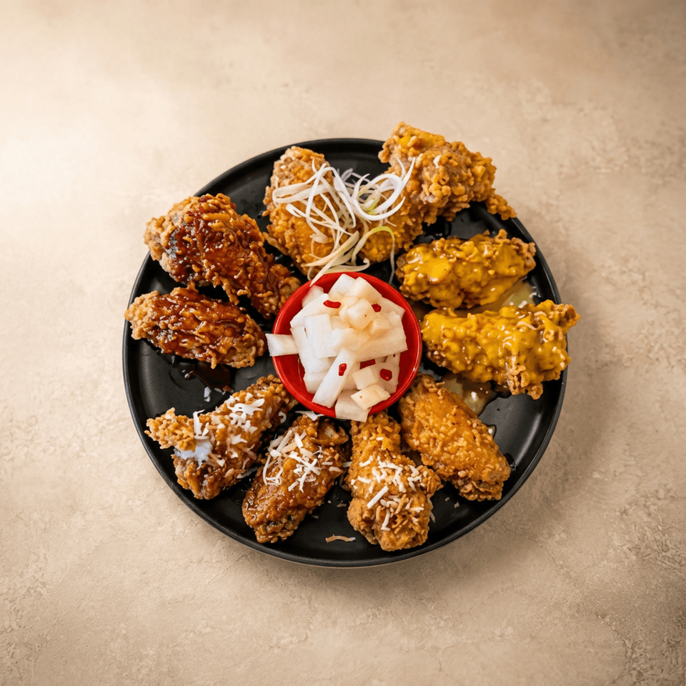 Assorted crispy fried chicken wings with varied sauces, shredded garnish, and pickled radish in a red bowl on a black plate.