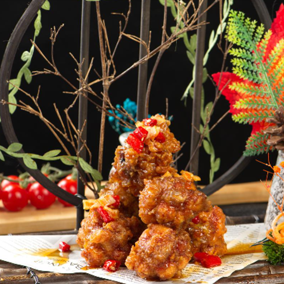 Glazed fried chicken pieces piled on paper, garnished with red berries, beside colorful autumn floral decor and tomatoes.