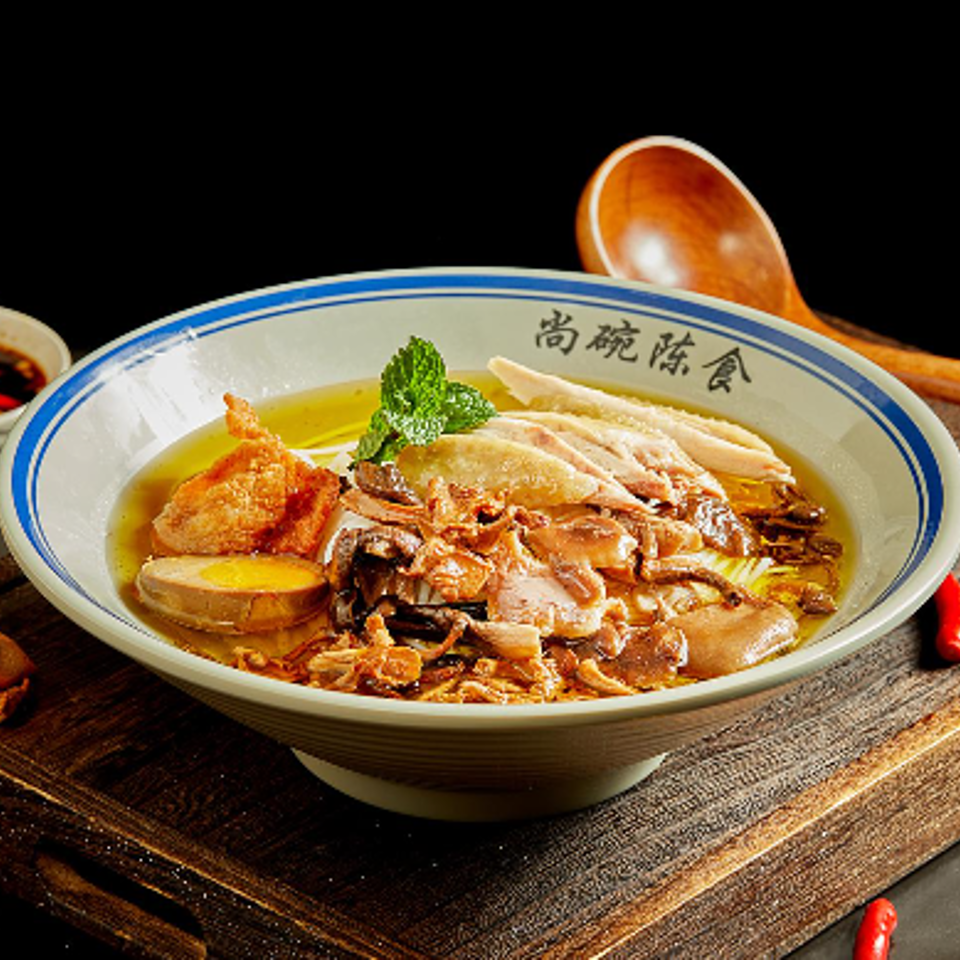 Chinese-style noodle soup topped with sliced pork, herbs, and vegetables in a ceramic bowl beside a wooden spoon.