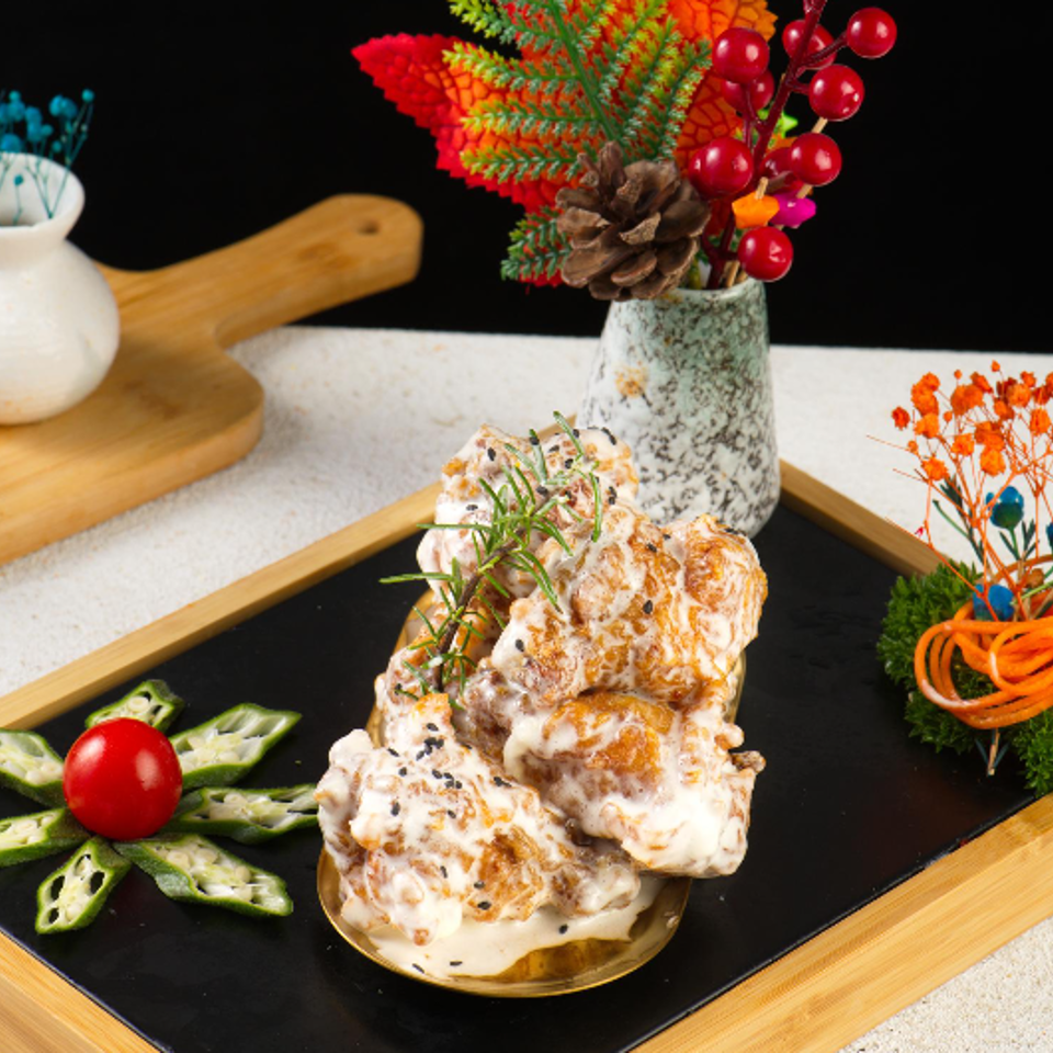 A richly browned, creamy savory dish garnished with herbs and sesame sits on a black serving board beside vegetable decorations.