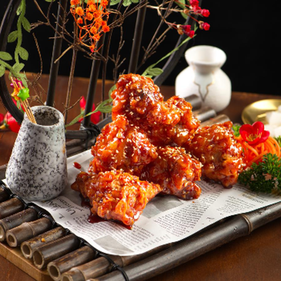 Glazed fried chicken piled on newspaper-lined bamboo mat, garnished with flowers and accompanied by dipping sauce.