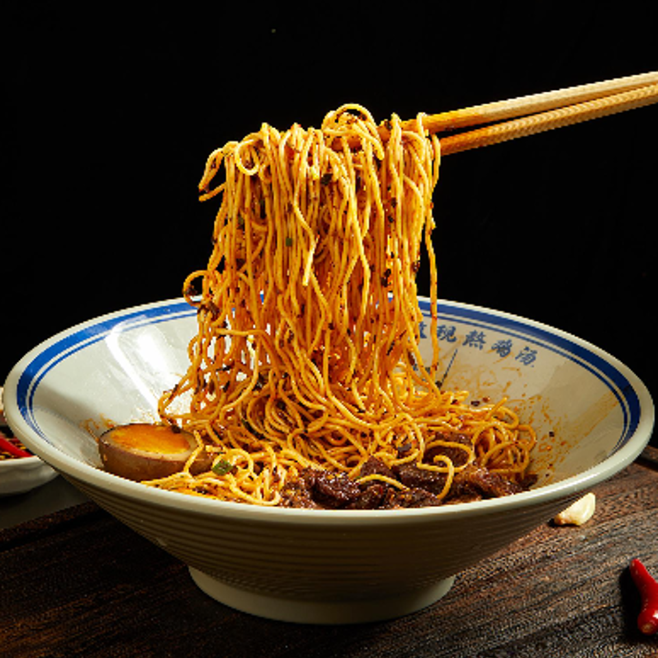 Chopsticks lift a tangled serving of saucy noodles from a ceramic bowl on a dark wooden table.